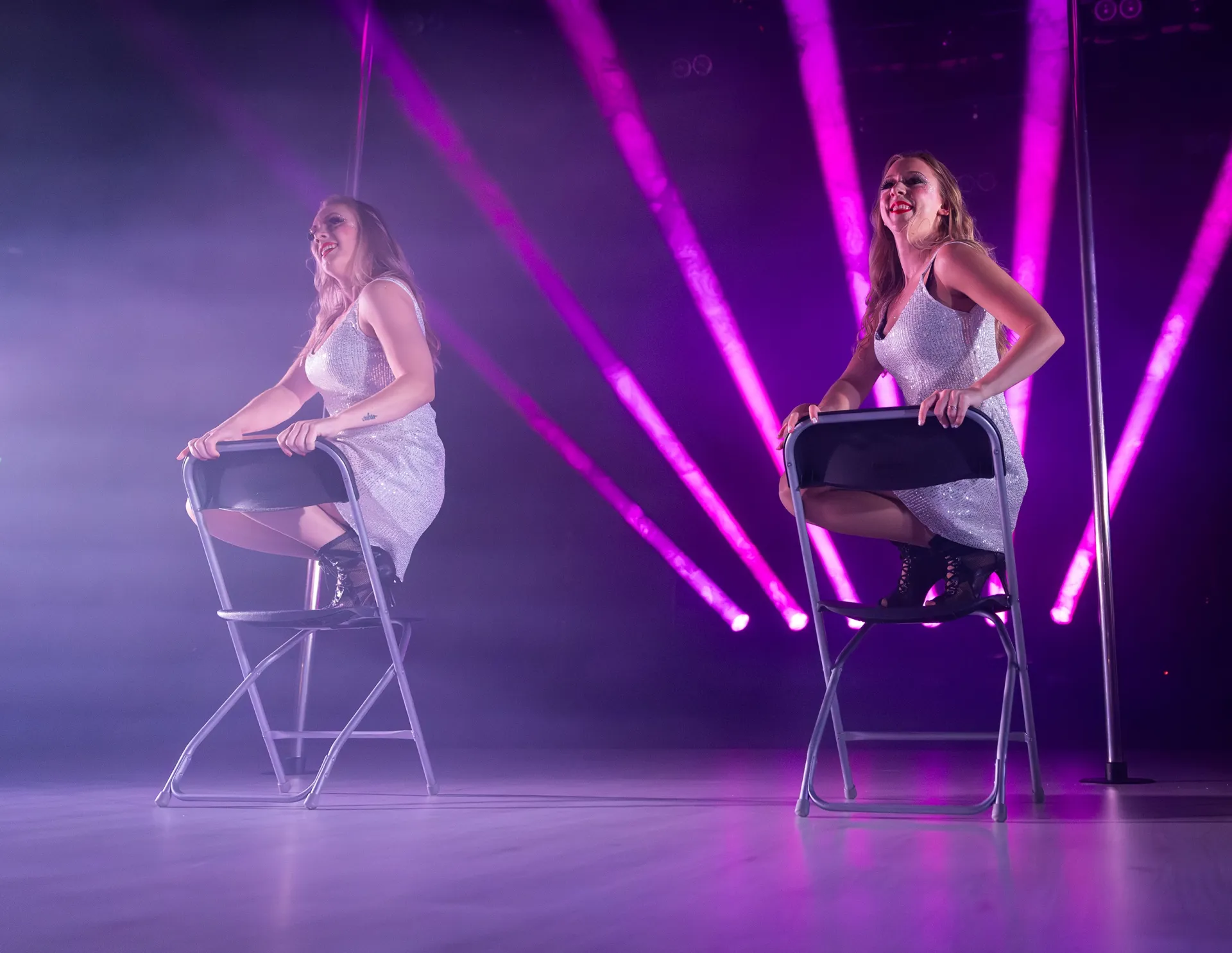 Två showartister uppträder på stolar i glittrande scenkostymer, upplysta av rosa och lila scenljus under ett elegant nummer.