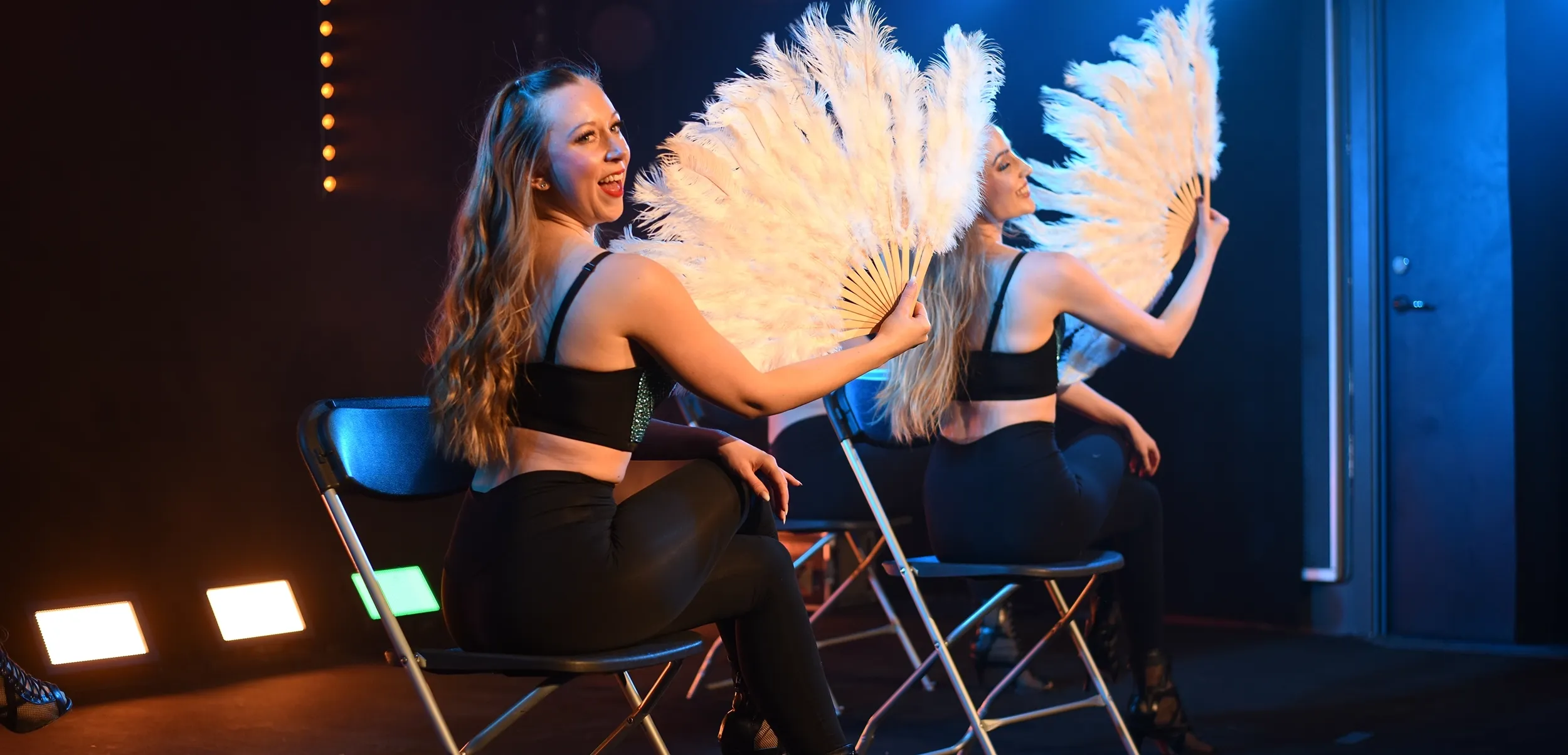 Ella och Sandra sitter på stolar i scenljus och showar med stora vita fjädervippor i klassisk burlesquestil.