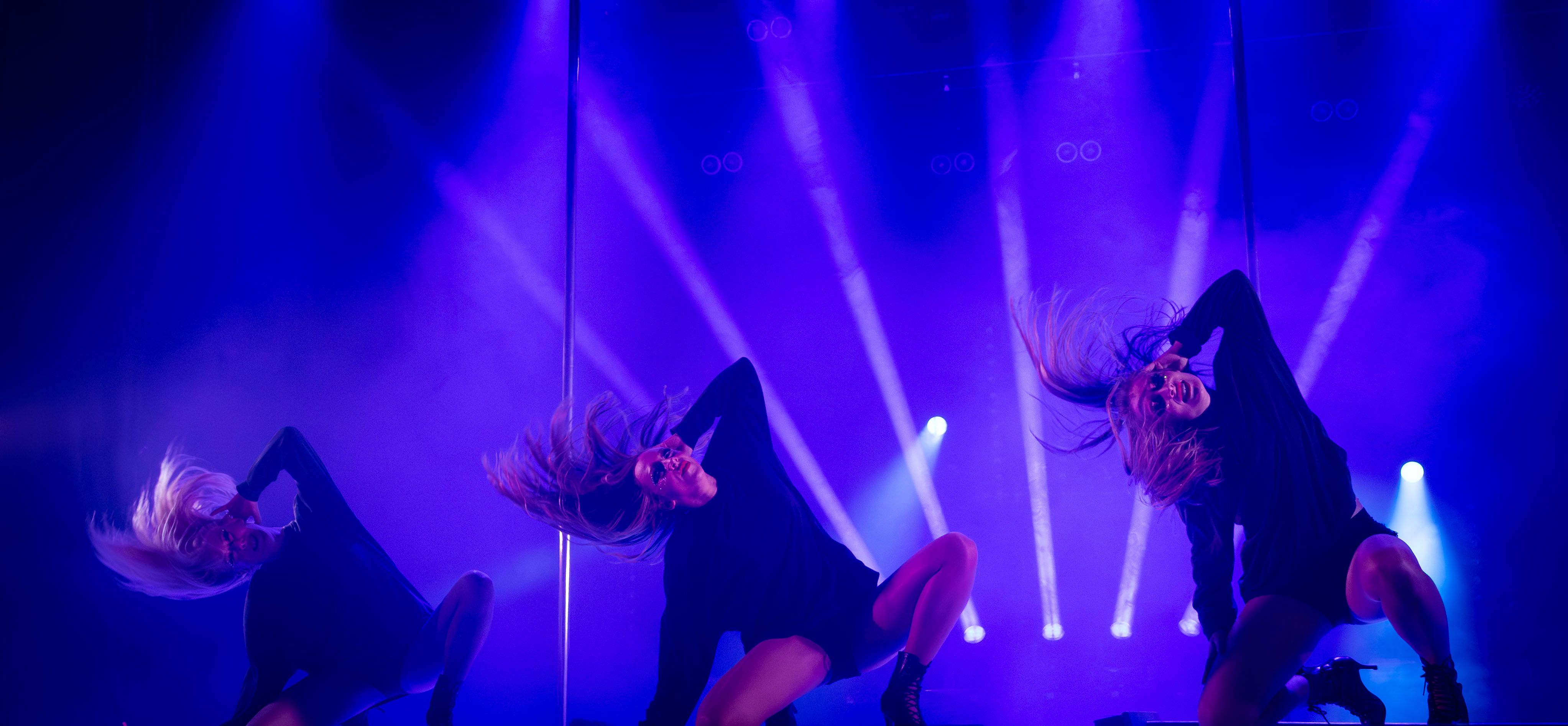 Tre dansare från Femme Fusion poserar i starkt blått scenljus under ett energiskt shownummer.