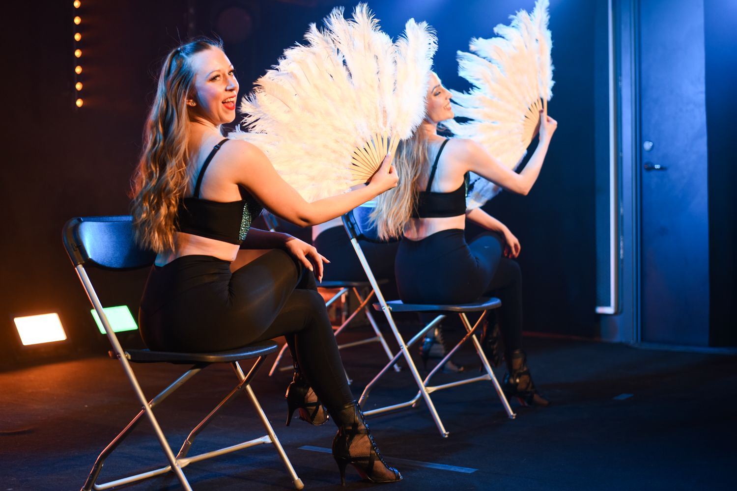 Två dansare sitter på stolar på scen och uppträder med stora vita fjäderfans i en elegant cabaret-inspirerad show.